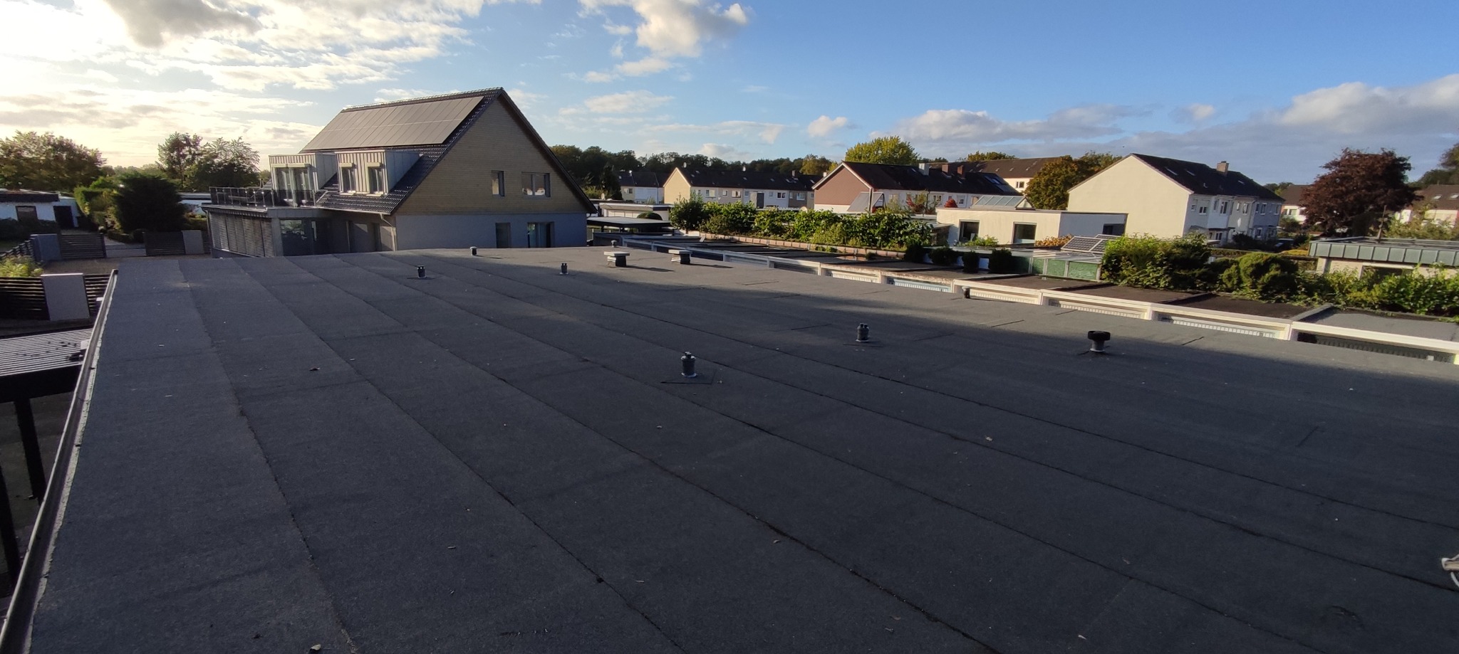 Czarny, płaski dach z papy termozgrzewalnej, widok z góry na tle zabudowy jednorodzinnej i błękitnego nieba z chmurami.