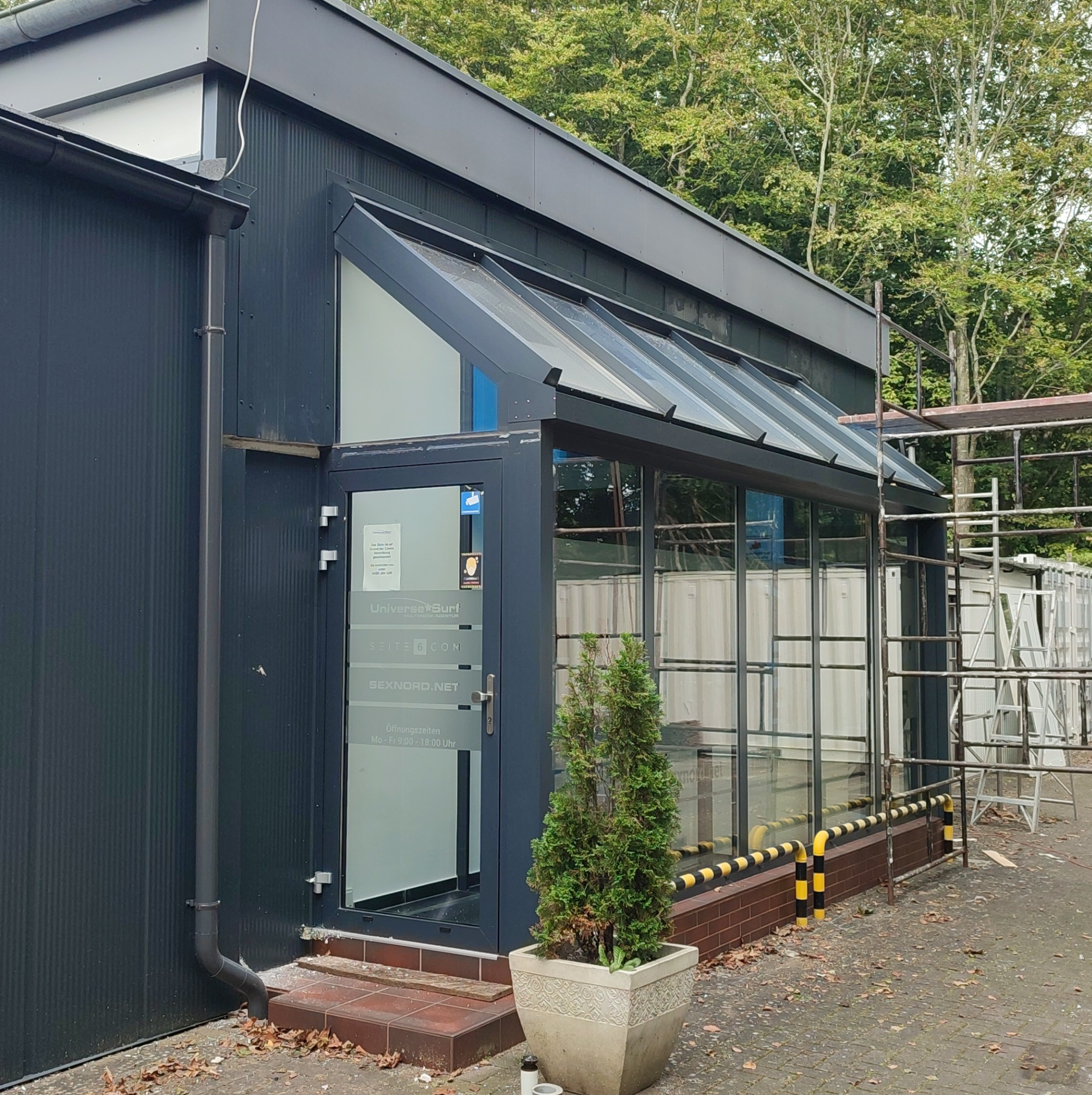 Nowoczesny budynek z przeszklonym wejściem i szarym dachem, widoczne rusztowania w tle, roślina w donicy przed wejściem.