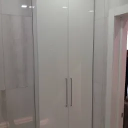 Biała, lakierowana szafa wnękowa z długimi, pionowymi uchwytami, wkomponowana w łazienkę wykończoną dużymi, jasnymi płytami imitującymi marmur, widoczny fragment białej ościeżnicy drzwi.