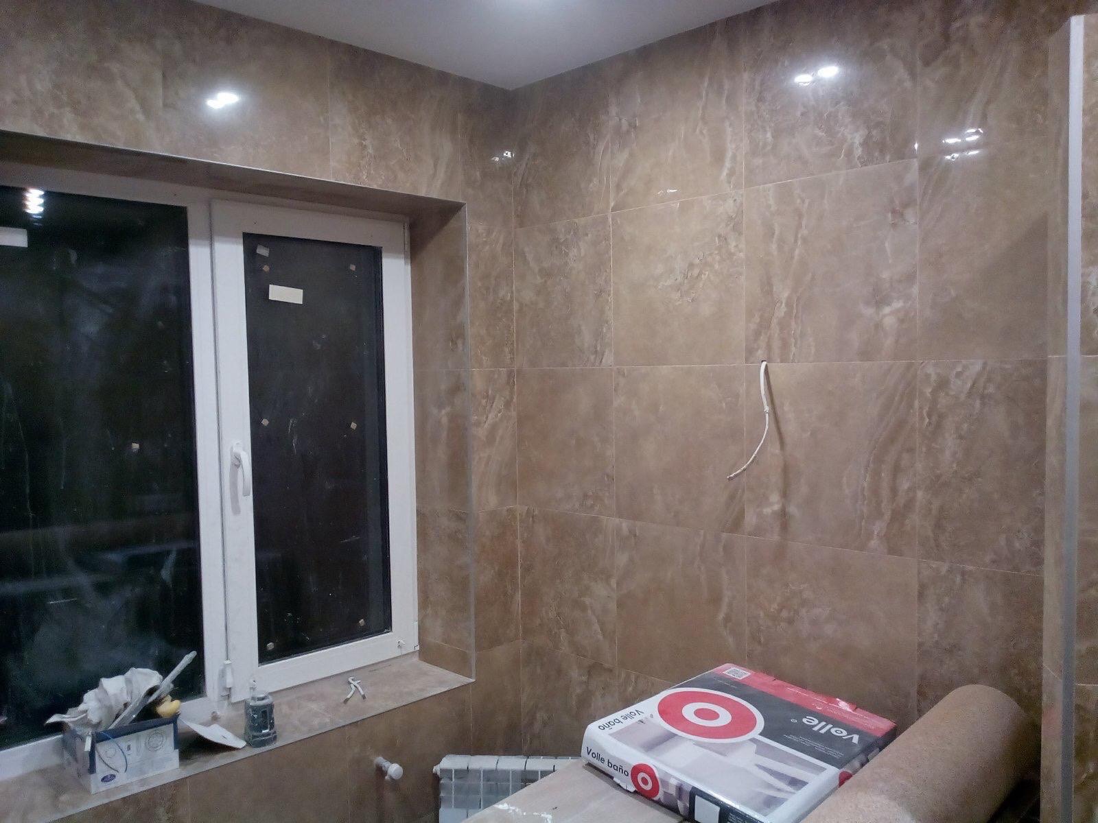 Ściany łazienki wyłożone brązowymi płytkami imitującymi marmur, widoczne okno z białymi ramami, narzędzia na parapecie i opakowanie płytek na podłodze, kabel wystający ze ściany.