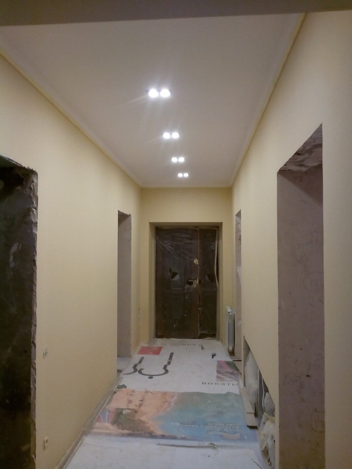 Korytarz w trakcie malowania na jasny odcień, podłoga zabezpieczona folią malarską z widocznymi napisami, sufit z rzędem podwójnych lamp halogenowych.