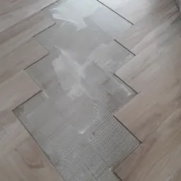 Układanie jasnych paneli podłogowych w jodełkę, widoczny klej na podłożu, etap prac.