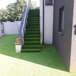 Zewnętrzne schody pokryte sztuczną trawą, z metalową barierką, prowadzące do drzwi na piętrze, obok domu z elewacją w dwóch odcieniach szarości.