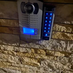 Szary panel domofonu z podświetlanymi na niebiesko przyciskami i kamerą, zamontowany na elewacji z kamienia dekoracyjnego.