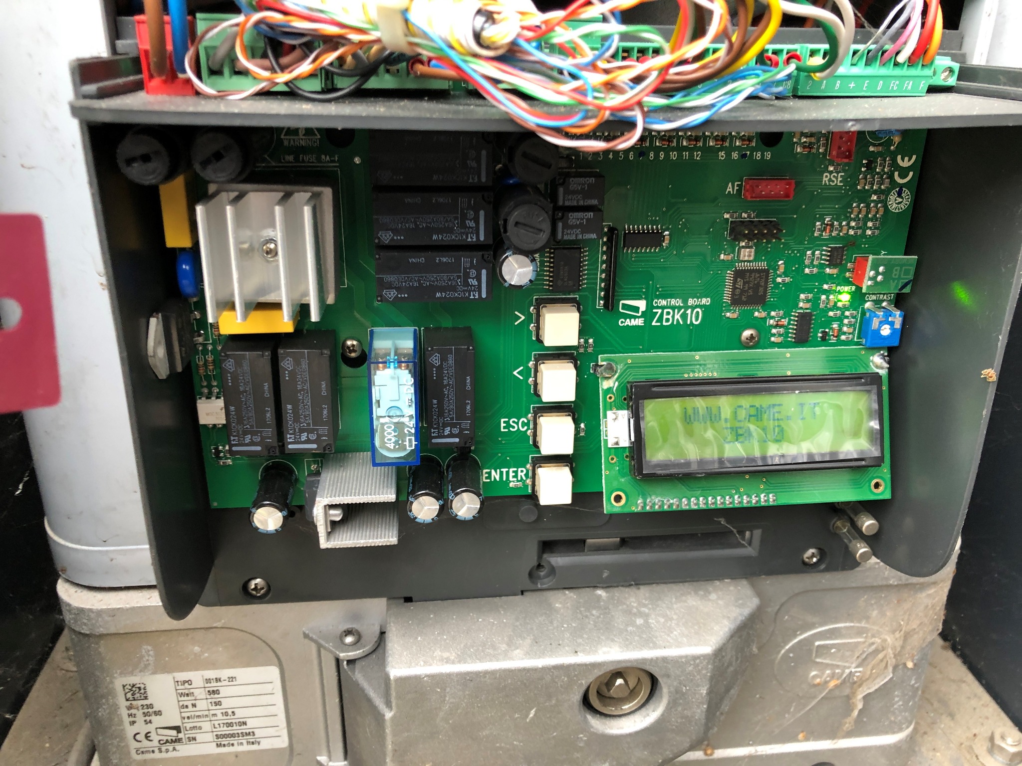 Otwarta obudowa centrali sterującej bramy CAME ZBK10, widoczna zielona płytka drukowana z elementami elektronicznymi, wyświetlaczem LCD pokazującym adres strony internetowej producenta i okablowaniem.