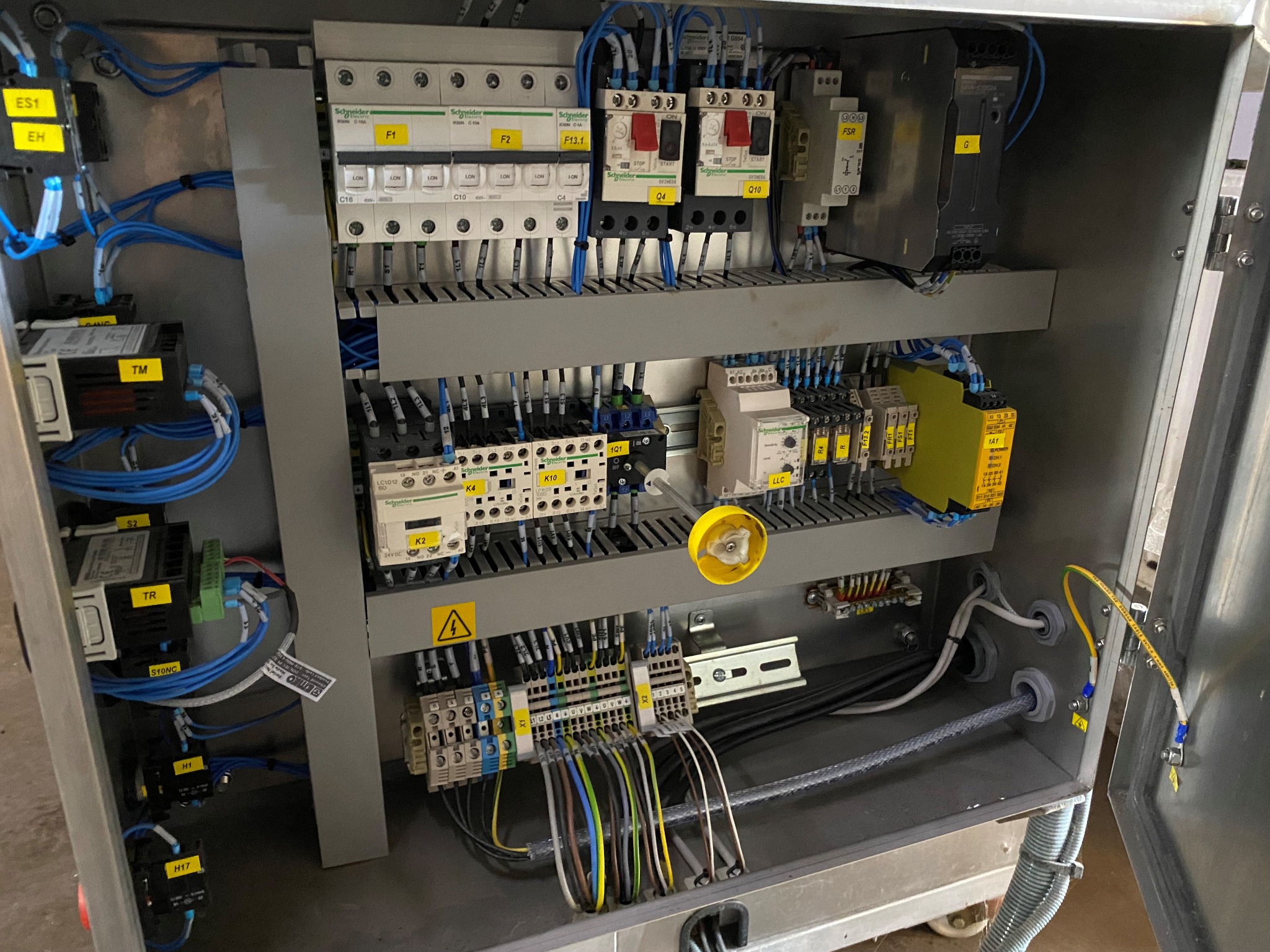 Otwarta szafa sterownicza z widocznymi elementami elektrycznymi, takimi jak przekaźniki, bezpieczniki, styczniki, listwy zaciskowe i okablowanie. Na elementach widoczne żółte etykiety...