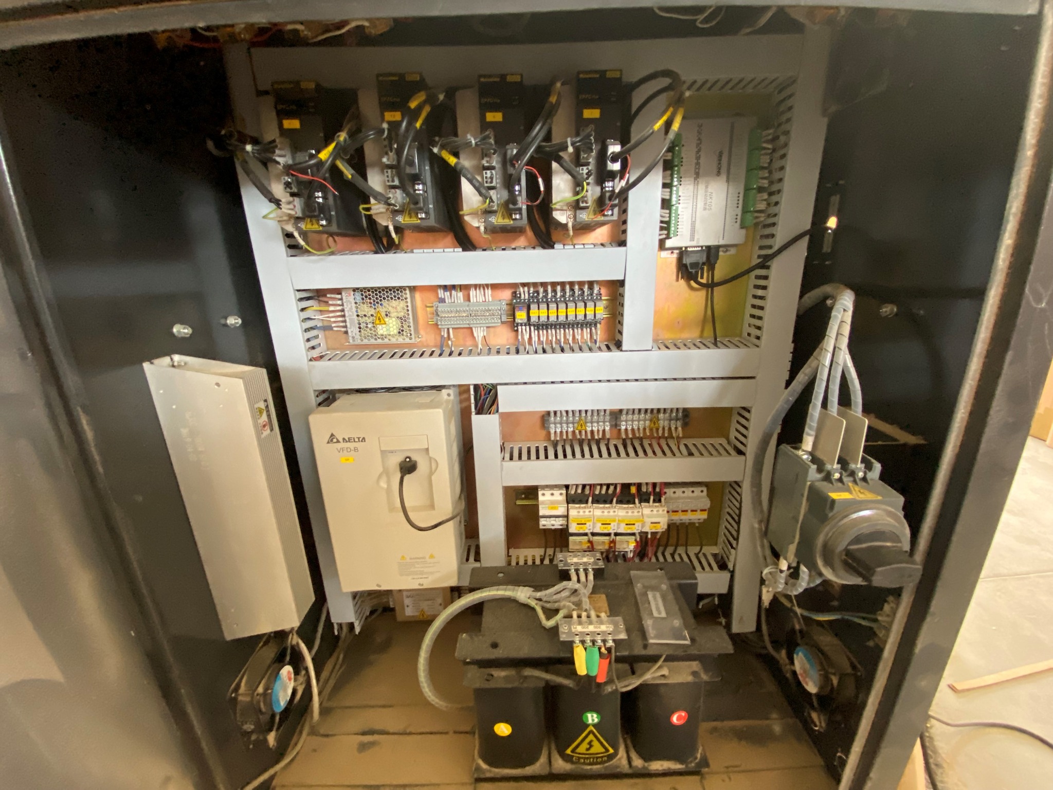 Wnętrze szafy sterowniczej z modułami, przewodami i transformatorami, widoczny panel Delta VFD-B oraz przełącznik obrotowy, całość w obudowie z czarną ramą.