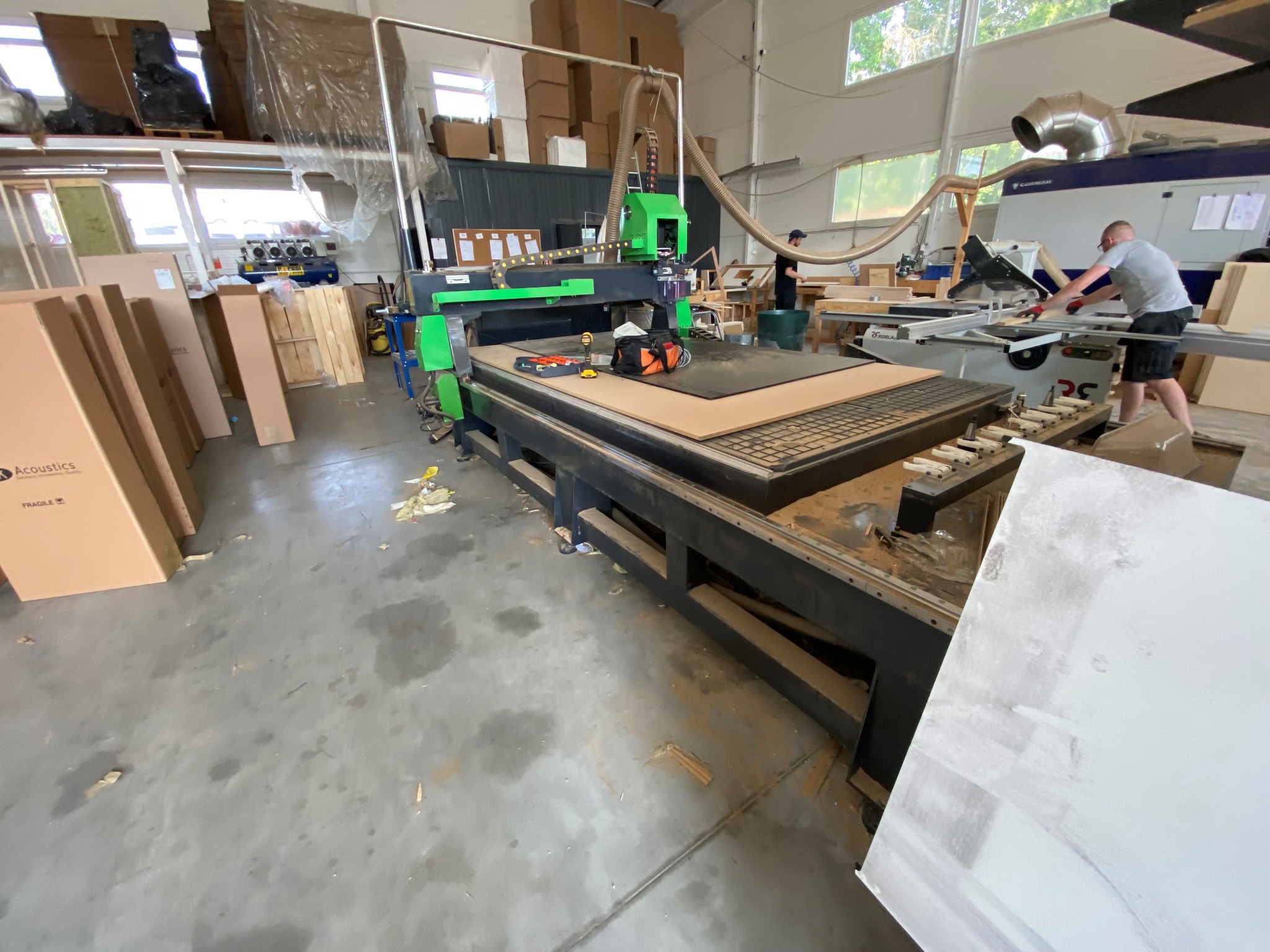 Szeroki kadr warsztatu z maszyną CNC w kolorze czarnym i jaskrawozielonym, obok której leżą pomarańczowe słuchawki ochronne i torba narzędziowa, a w tle widać mężczyznę obsługującego piłę stołową.