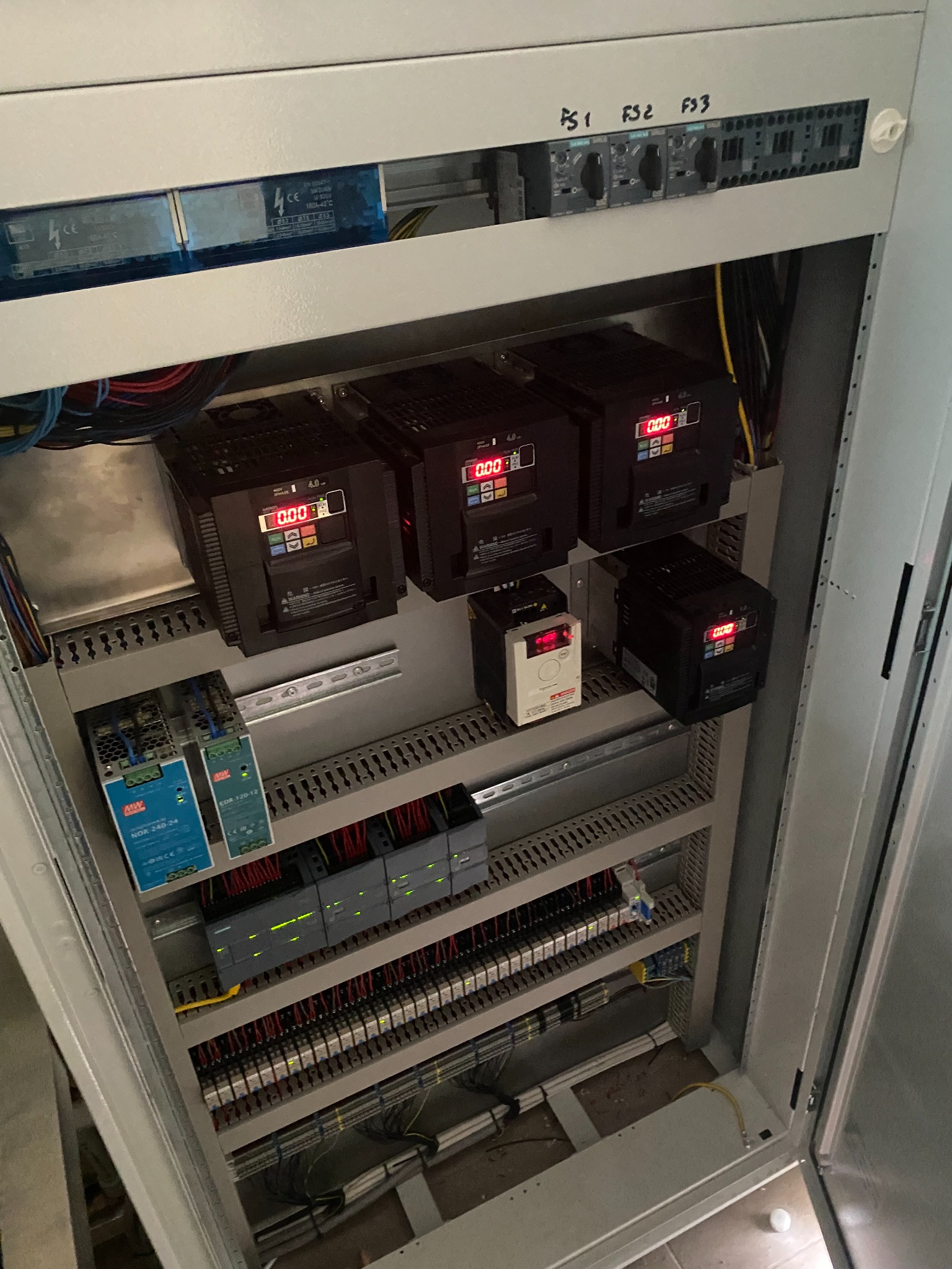 Otwarta szafa sterownicza z widocznymi modułami PLC Siemens, zasilaczami impulsowymi MW Power, falownikami z wyświetlaczami LED i licznymi połączeniami kablowymi, zasilanie trójfazowe opisane...