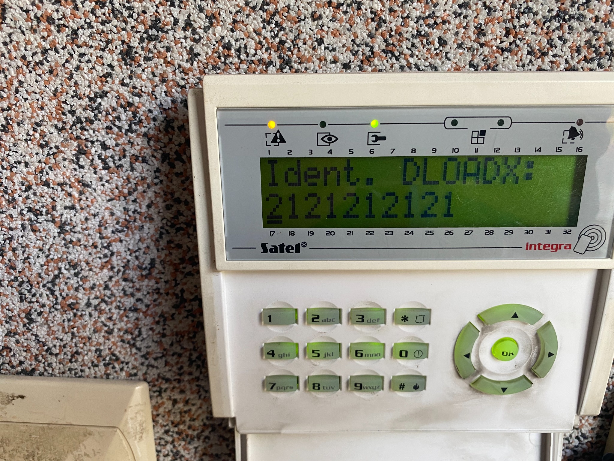 Panel sterowania alarmem domowym firmy Satel Integra z wyświetlonym komunikatem 'Ident. DLOADX: 2121212121', zamontowany na ścianie o fakturze baranka, widoczne diody sygnalizacyjne i klawiatura...