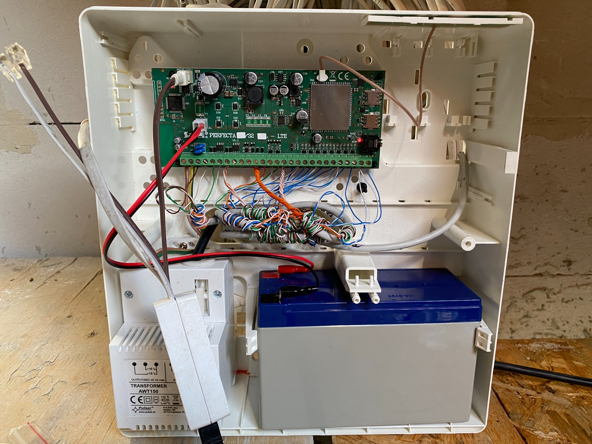 Wnętrze skrzynki alarmowej z płytą główną Perfecta LTE, akumulatorem i transformatorem Pulsar AWT150, widoczne liczne przewody i złącza.
