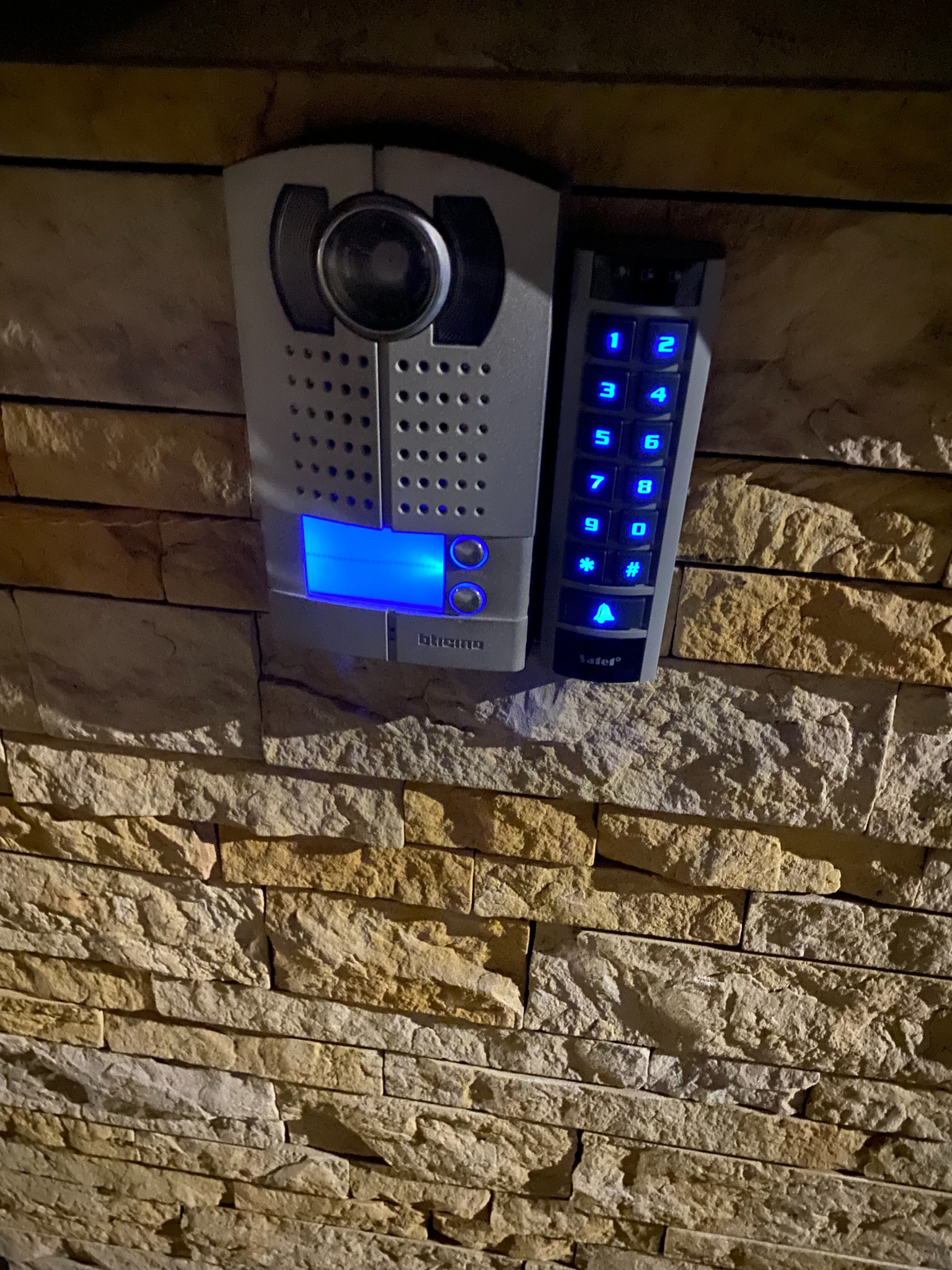 Szary panel domofonu z podświetlanymi na niebiesko przyciskami i kamerą, zamontowany na elewacji z kamienia dekoracyjnego.