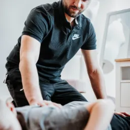 Mężczyzna w czarnej koszulce polo z logo Nike, klęczy nad pacjentem leżącym na podłodze i wykonuje masaż pleców, skupiony wzrok.