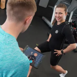Kobieta w czarnym stroju EMS wykonuje wykrok, uśmiechając się do kamery, podczas gdy trener monitoruje parametry na tablecie w siłowni.