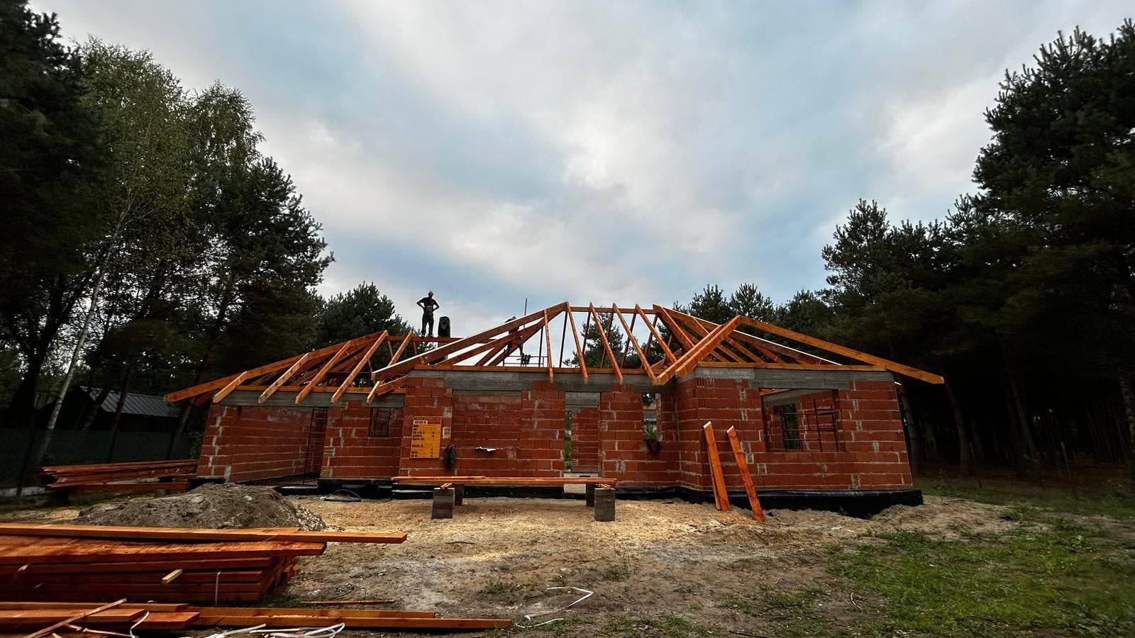 Budowa domu jednorodzinnego w stanie surowym otwartym: murowane ściany z czerwonej cegły, drewniana konstrukcja dachu w trakcie montażu, widoczna sylwetka pracownika na dachu, stos drewna...
