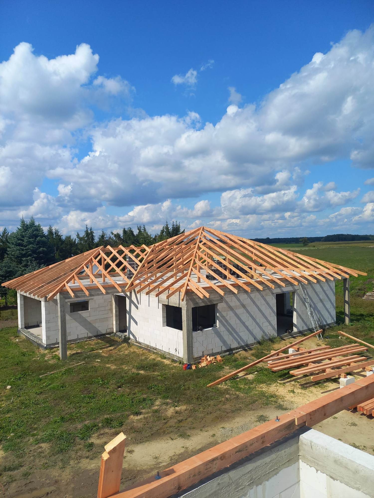 Budowa domu jednorodzinnego z widoczną konstrukcją drewnianą dachu na tle błękitnego nieba z chmurami.