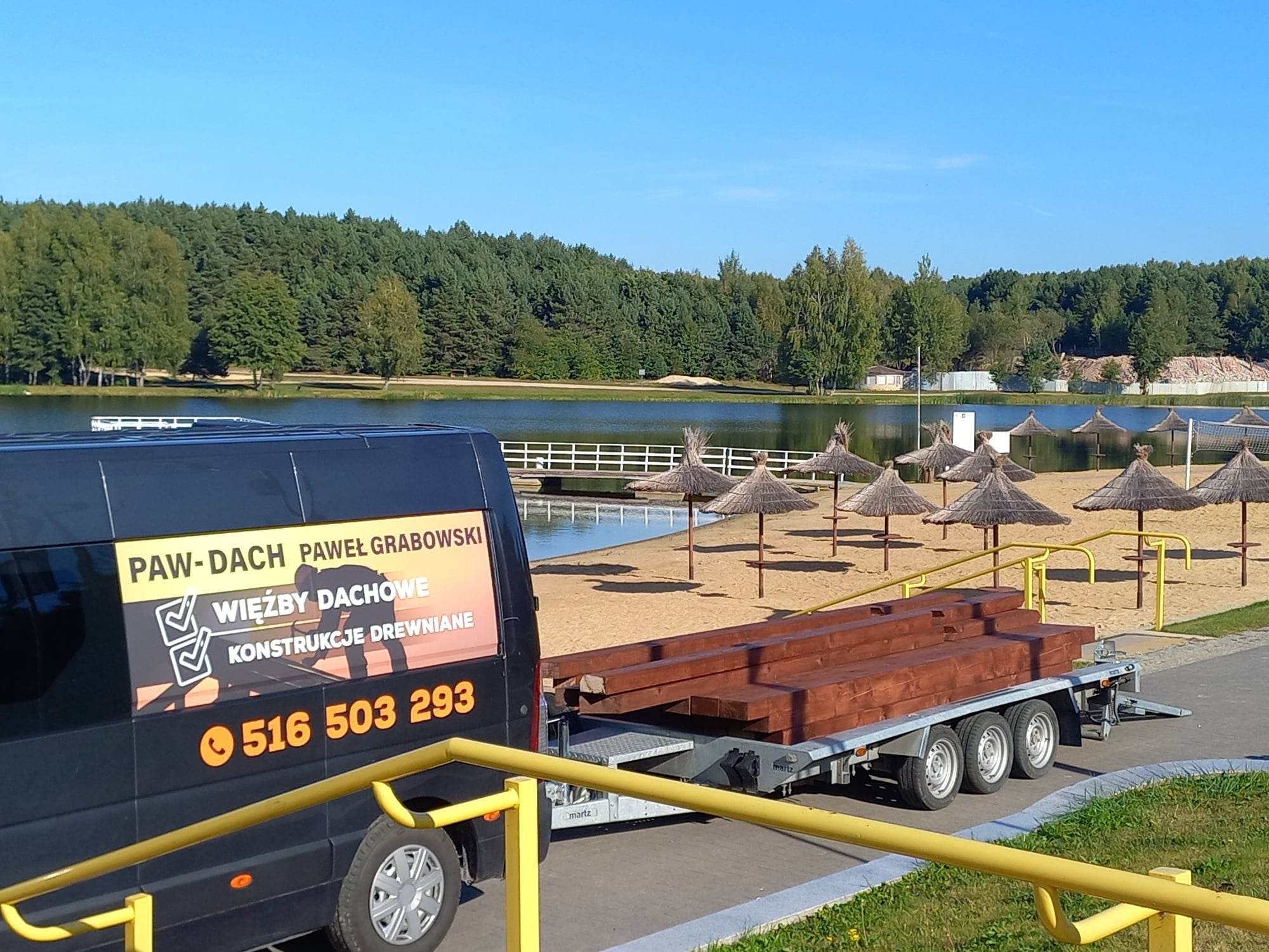 Ciemny bus firmy Paw-Dach Pawel Grabowski z reklamą więźby dachowej i numerem telefonu, ciągnący przyczepę z brązowymi belkami drewnianymi na tle jeziora z plażą i parasolami.
