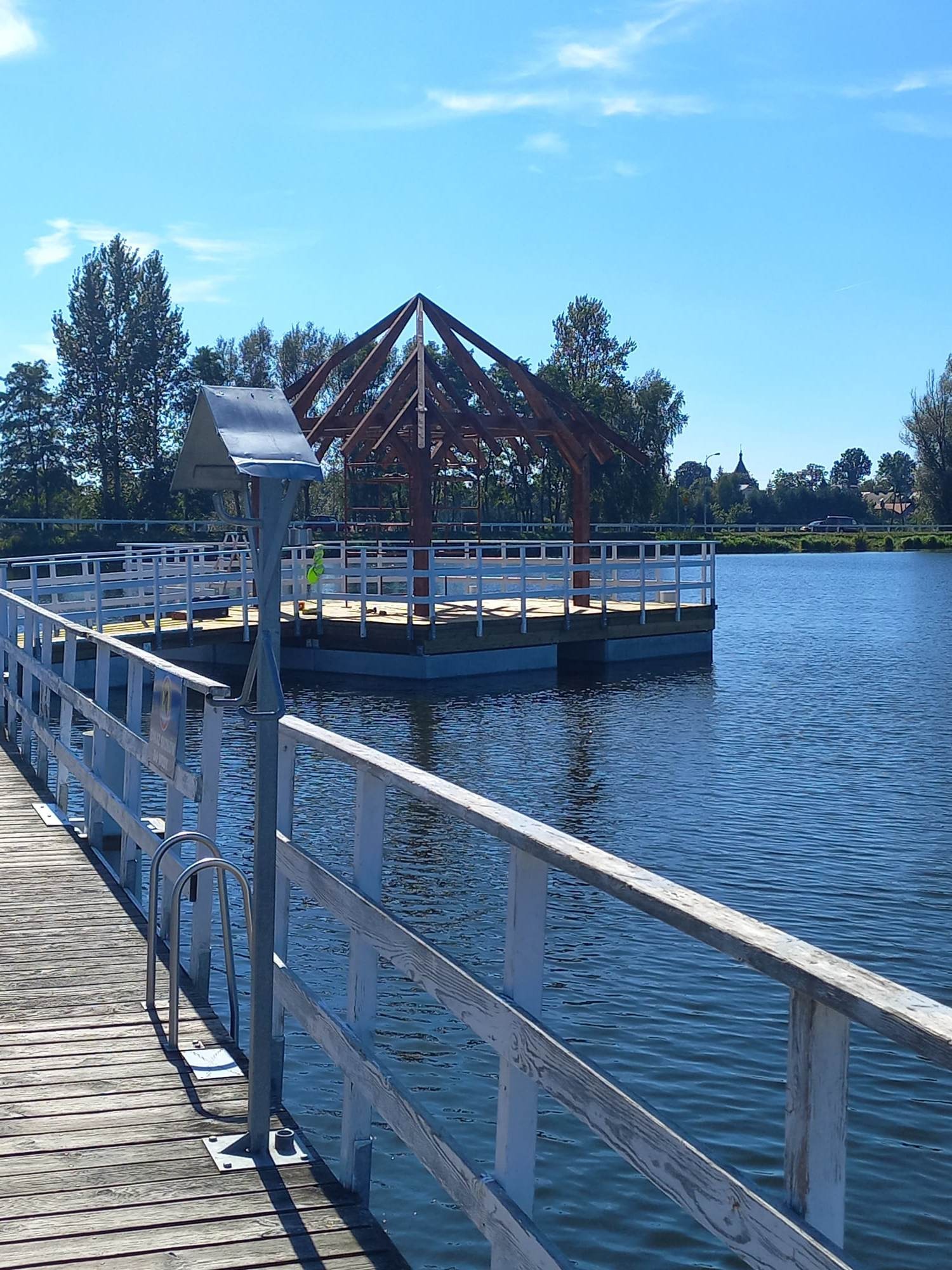 Konstrukcja drewnianej altany z więźbą dachową na platformie na wodzie, widok z drewnianego pomostu, błękitne niebo w tle.