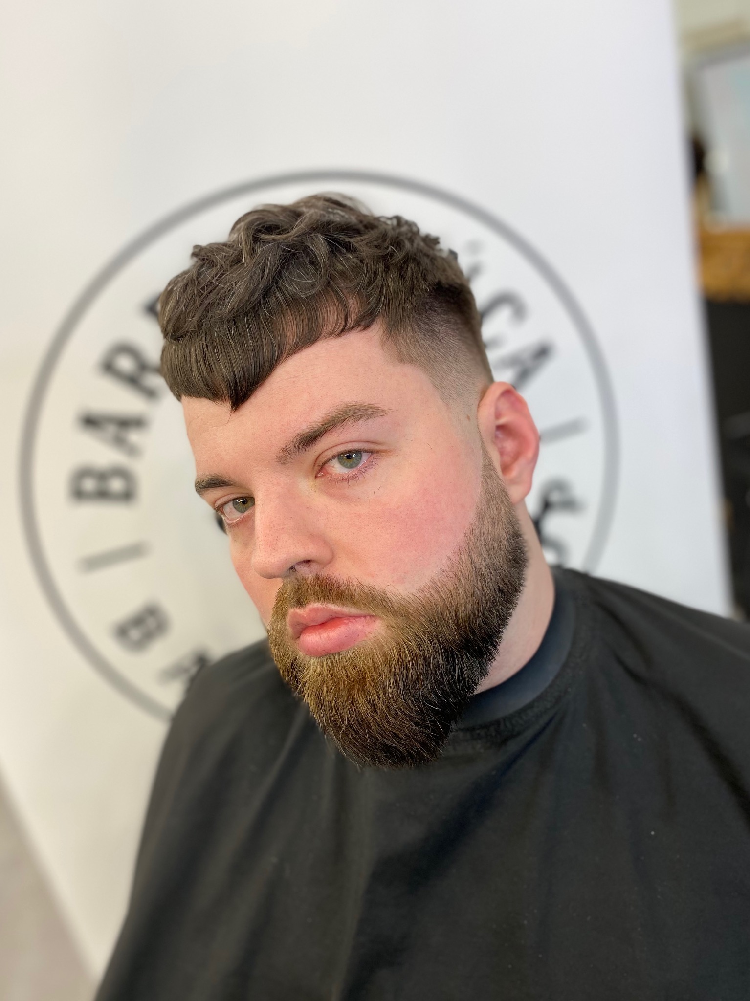 Mężczyzna z krótką, teksturowaną fryzurą i przystrzyżoną brodą, pozujący na tle ściany z logo salonu.
