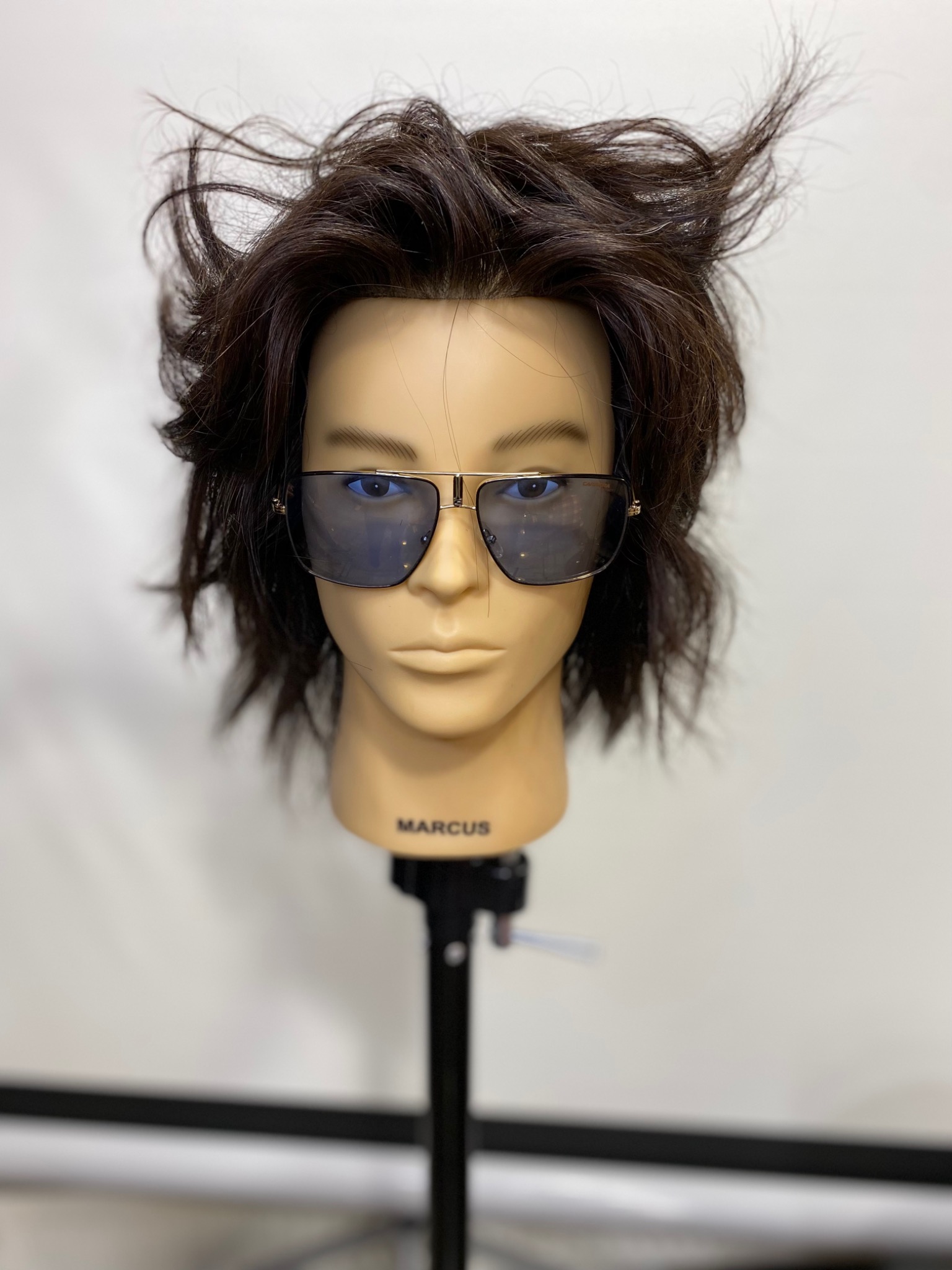 Manekin z ciemnymi, potarganymi włosami i okularami przeciwsłonecznymi, umieszczony na czarnym stojaku z napisem 'MARCUS'.