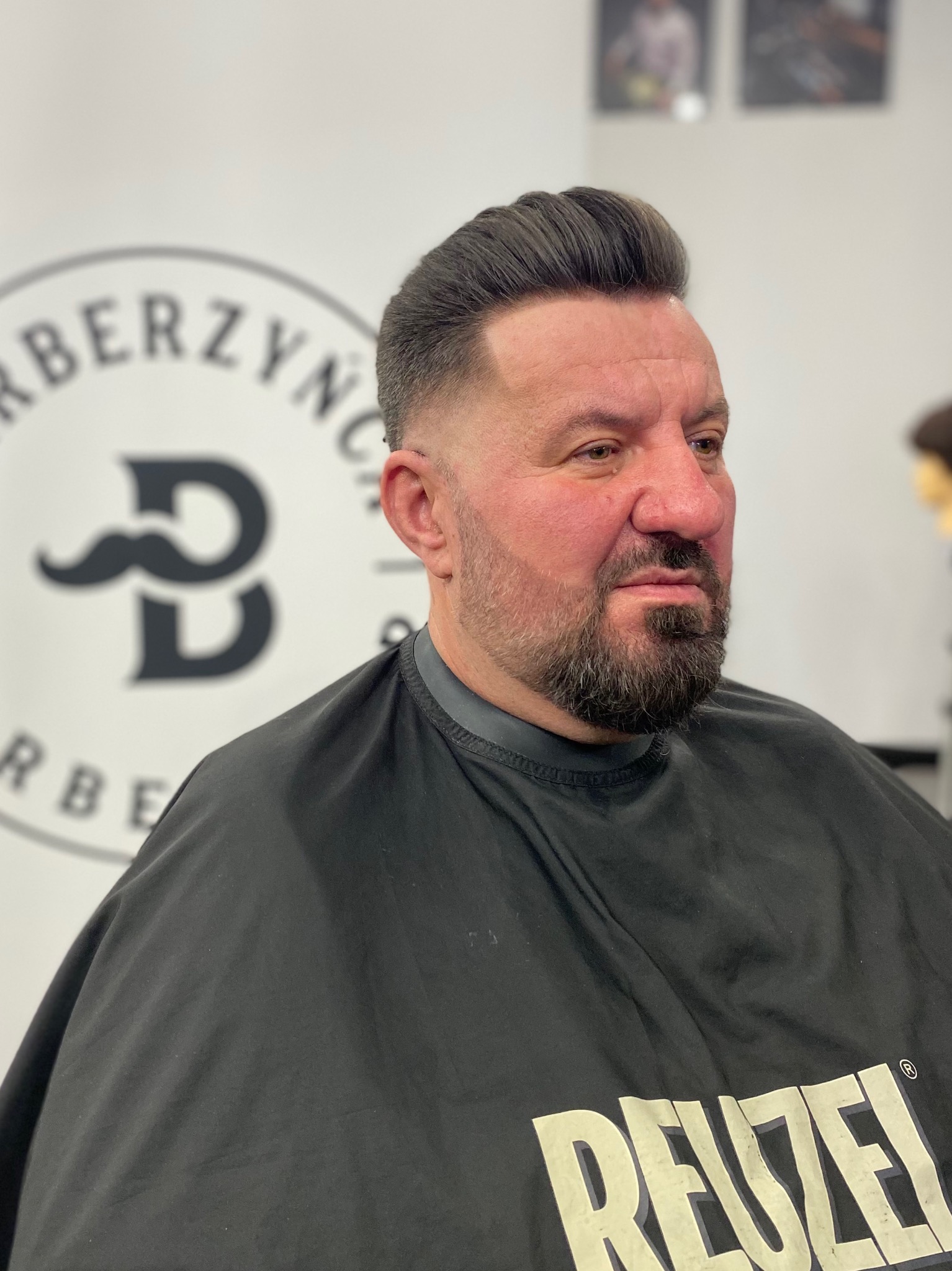 Mężczyzna po strzyżeniu z wycieniowanymi bokami i starannie ułożoną górą, ubrany w pelerynę z napisem 'REUZEL', na tle logo barber shopu z wąsami.