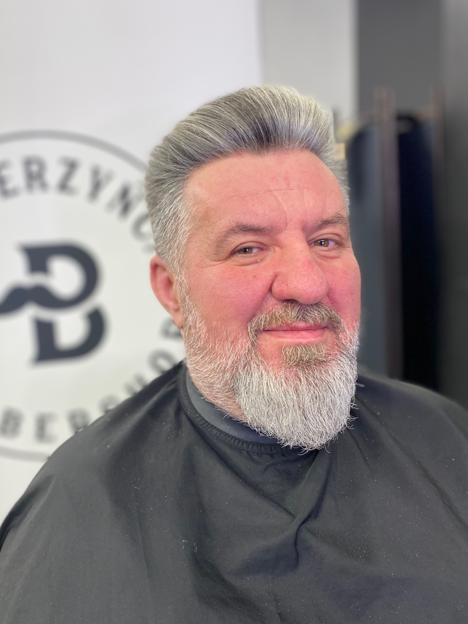 Mężczyzna z siwą brodą i włosami po strzyżeniu w salonie fryzjerskim, ubrany w czarną pelerynę fryzjerską, uśmiecha się do kamery na tle logo salonu.