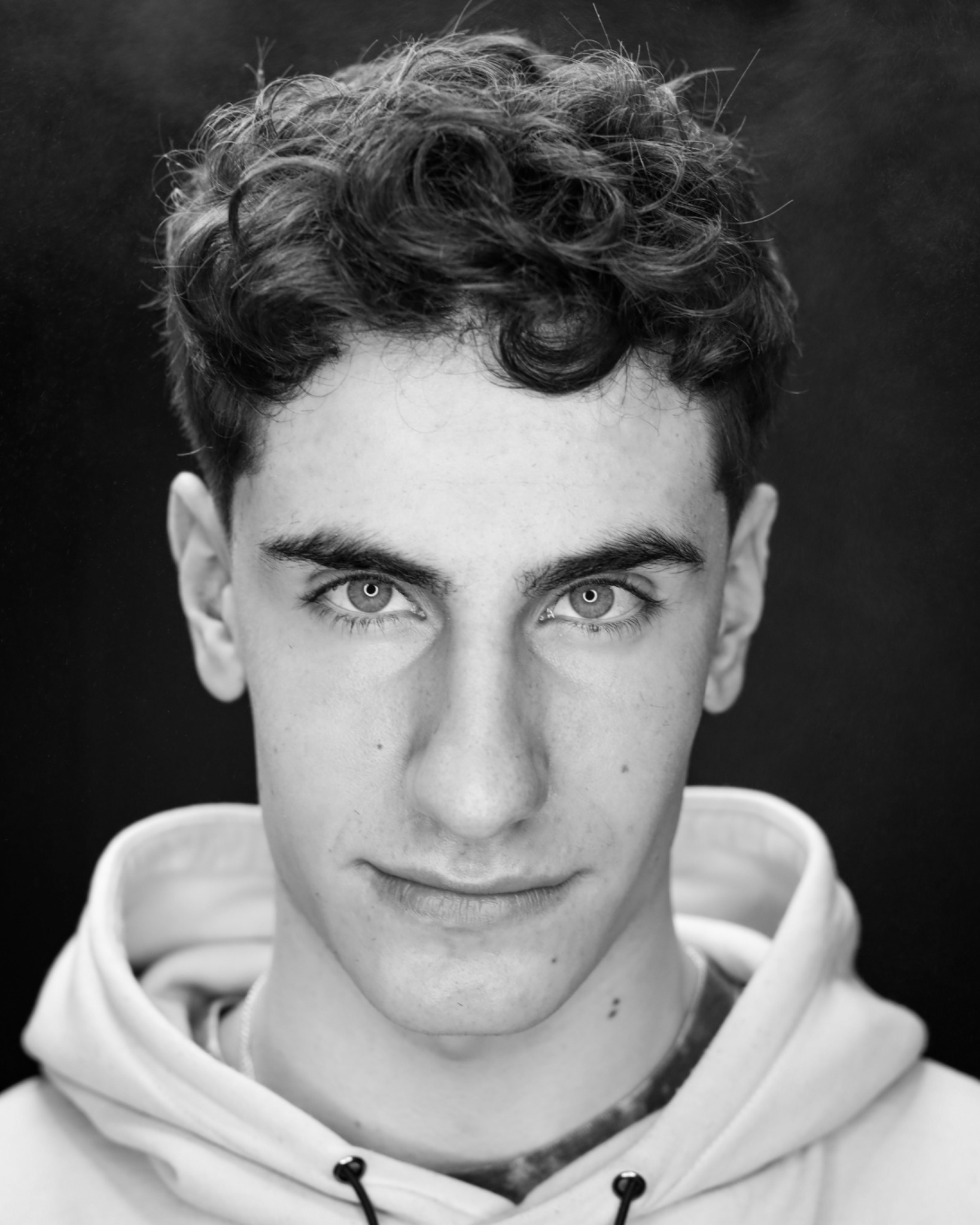 Portret młodego mężczyzny z kręconymi włosami w czerni i bieli, ubrany w bluzę z kapturem, na ciemnym tle.