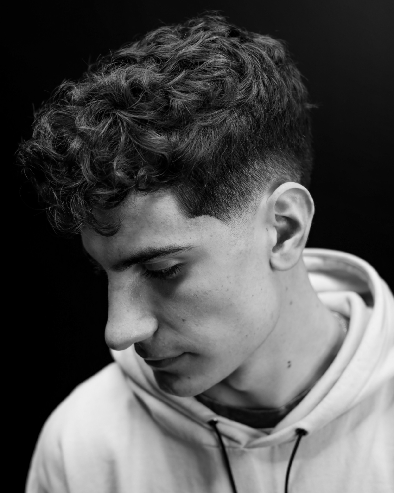 Portret w czerni i bieli młodego mężczyzny z krótkimi, kręconymi włosami, spuszczonym wzrokiem i delikatnym cieniowaniem fryzury.