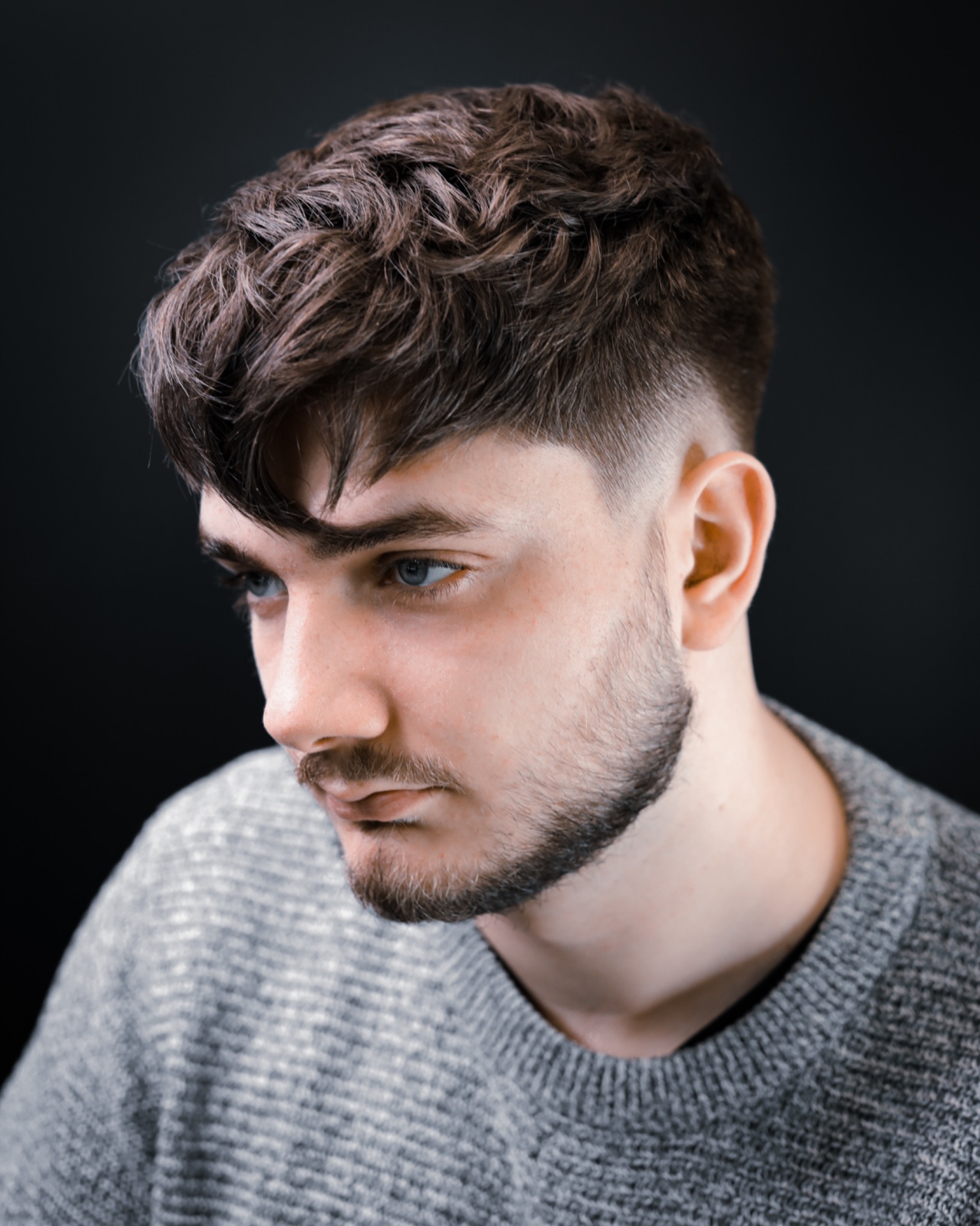 Portret młodego mężczyzny z modnym, cieniowanym cięciem włosów i krótką brodą, na ciemnym tle.