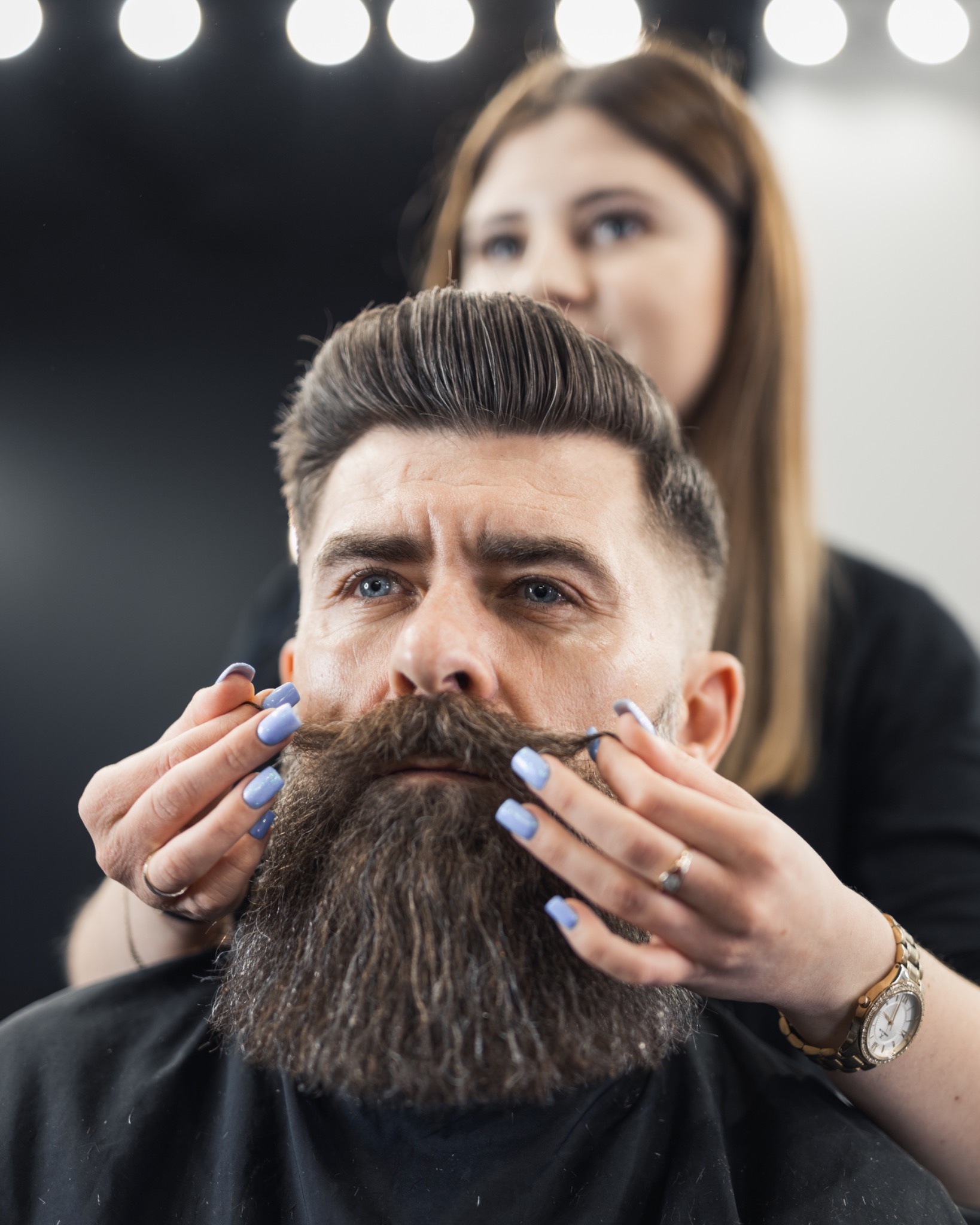 Mężczyzna z brodą i wąsami w trakcie stylizacji przez fryzjerkę, która poprawia mu wąsy dłońmi z jasnoniebieskim manicure, w tle oświetlenie salonu.
