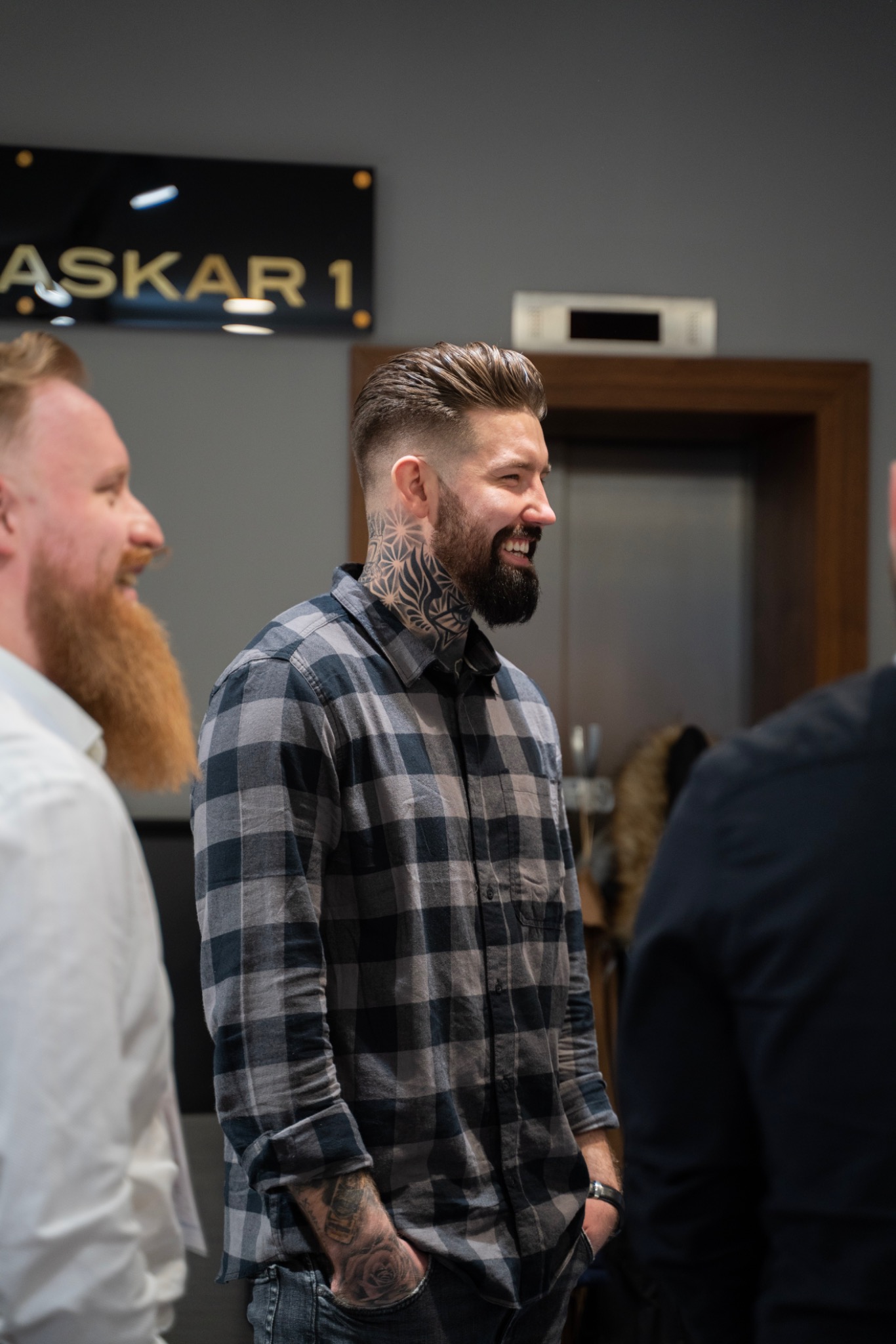 Mężczyzna z modną fryzurą, wytatuowaną szyją i brodą, uśmiecha się w salonie. W tle widoczny drugi mężczyzna z rudą brodą oraz tabliczka z napisem ASKAR 1.