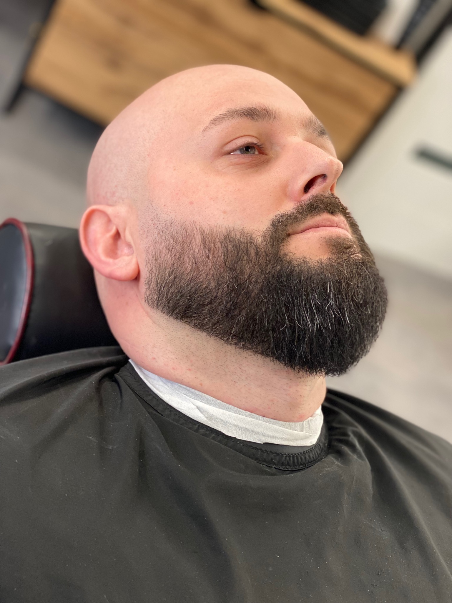 Mężczyzna z ogoloną głową i wystylizowaną brodą, widziany z dołu, ubrany w czarną pelerynę ochronną w salonie.