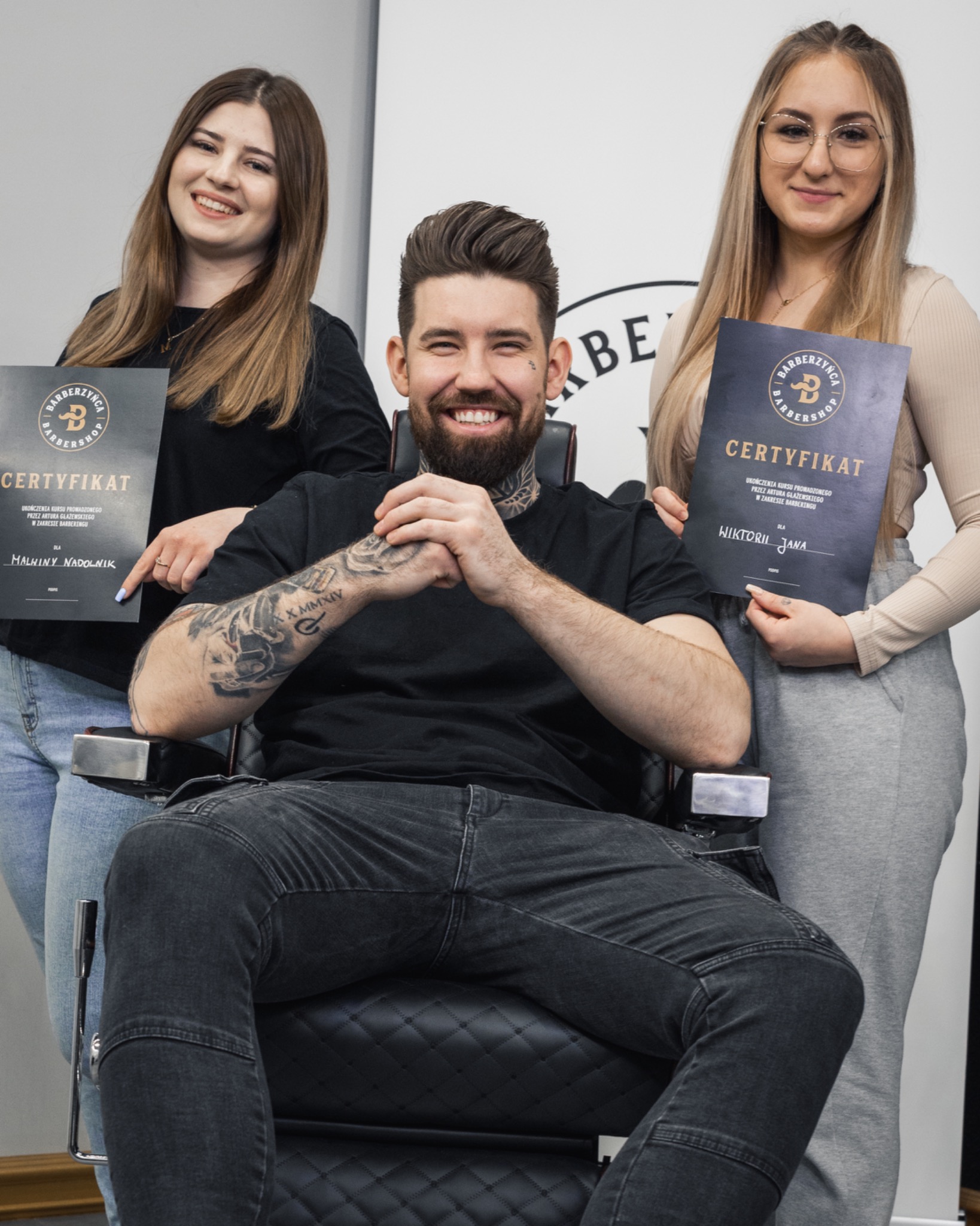 Uśmiechnięty brodaty mężczyzna z tatuażami siedzi w fotelu barberskim, otoczony przez dwie kobiety trzymające certyfikaty ukończenia kursu barberingu.