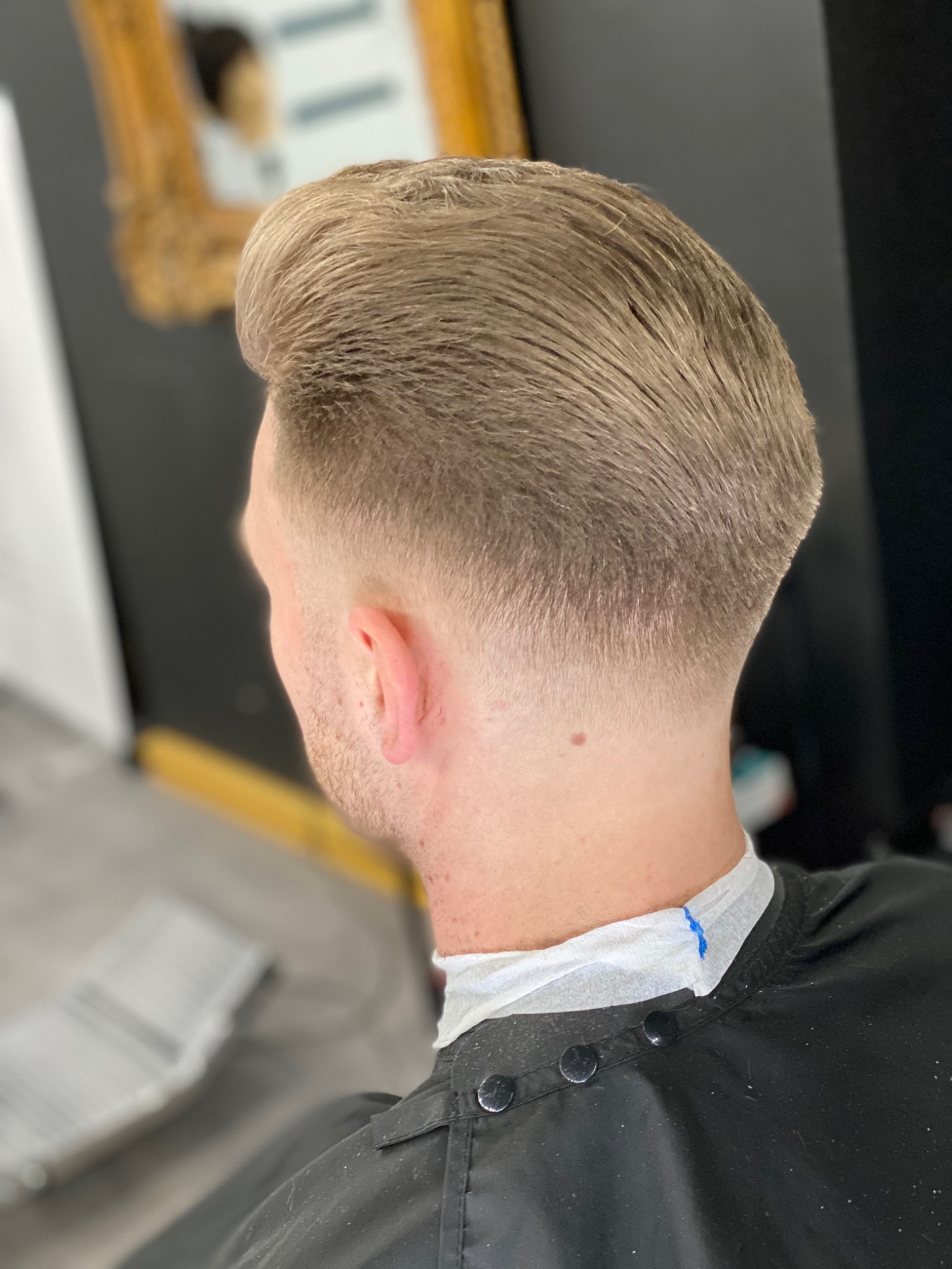 Męska fryzura z krótkimi bokami i zaczesaną do góry, blond czupryną, widok z tyłu na kark z pieprzykiem, peleryna ochronna z widocznymi guzikami i śladami farby.