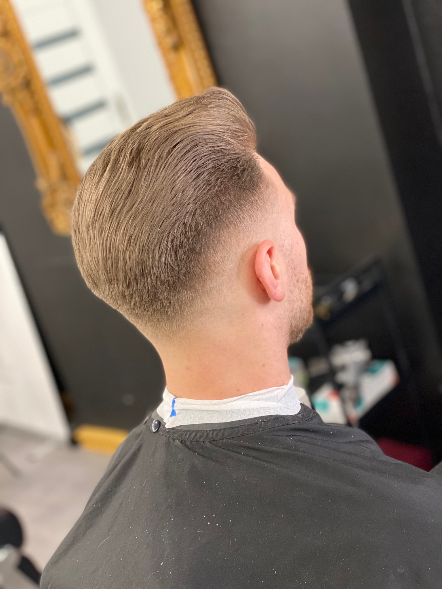 Męska fryzura z wycieniowanymi bokami i dłuższym, zaczesanym do góry włosem na górze głowy, widoczna z tyłu, z czarną peleryną ochronną.