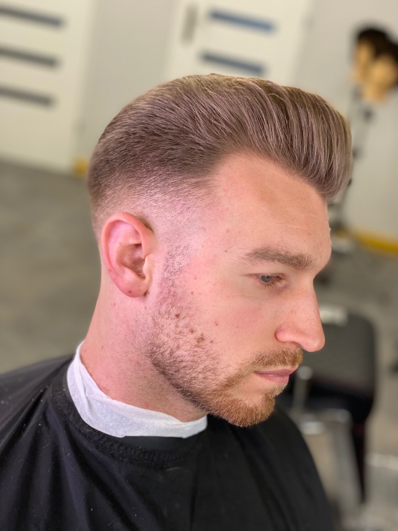 Męska fryzura typu pompadour z precyzyjnym cieniowaniem, widoczna z profilu, z delikatnym zarostem i peleryną fryzjerską.