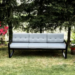 Metalowy zestaw ogrodowy z szarymi poduszkami, składający się z sofy i dwóch foteli, ustawiony na trawniku w otoczeniu drzew i kwiatów.