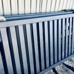 6 - Metalowa brama wjazdowa z pionowymi szczeblami, świeżo pomalowana na szaro, leżąca na podłożu z resztkami farby.