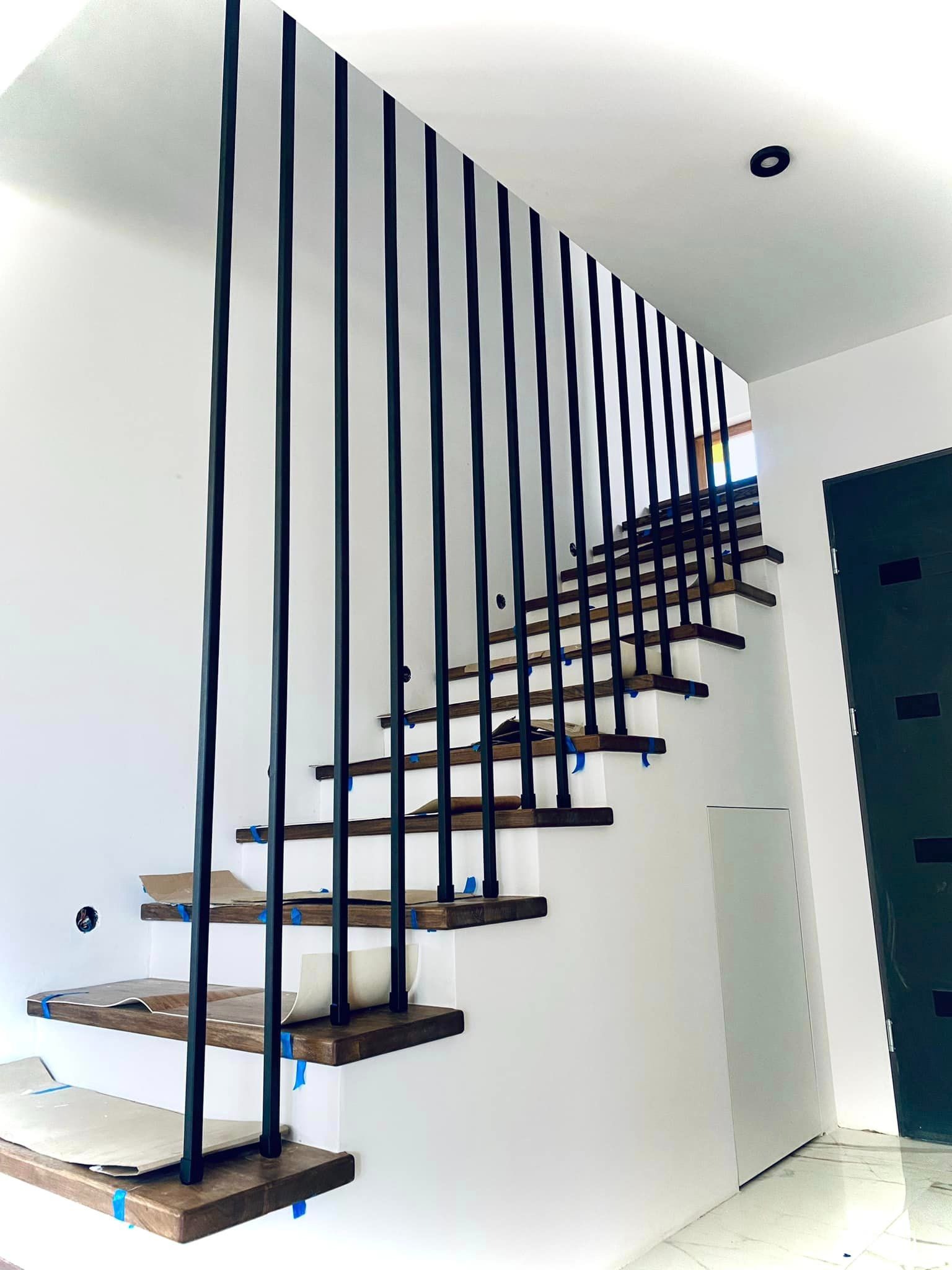 Nowoczesne schody z drewnianymi stopniami i czarnymi, pionowymi balustradami, zabezpieczone taśmą malarską i kartonem podczas prac wykończeniowych.