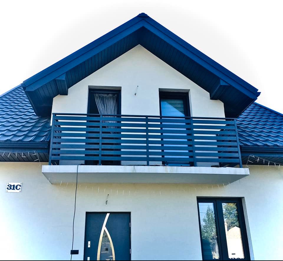 Dwupiętrowy dom z balkonem z czarną, poziomą balustradą, ciemnogranatowym dachem i jasną elewacją. Widoczne okna z firankami i numer domu 31C.
