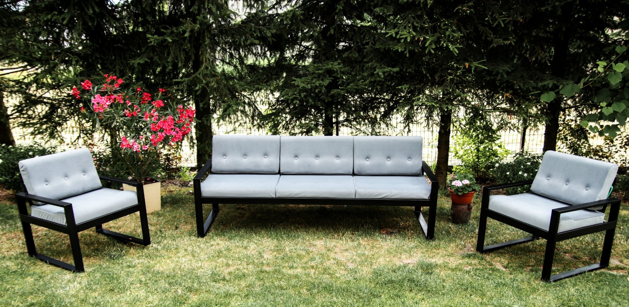 Metalowy zestaw ogrodowy z szarymi poduszkami, składający się z sofy i dwóch foteli, ustawiony na trawniku w otoczeniu drzew i kwiatów.