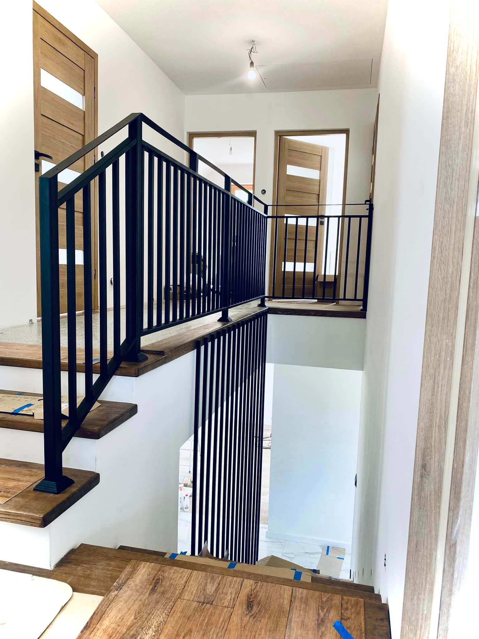Nowoczesna, czarna balustrada schodowa z pionowymi szczeblami, drewniane stopnie w trakcie montażu, widoczne zabezpieczenia kartonem, jasne wnętrze z drewnianymi drzwiami.