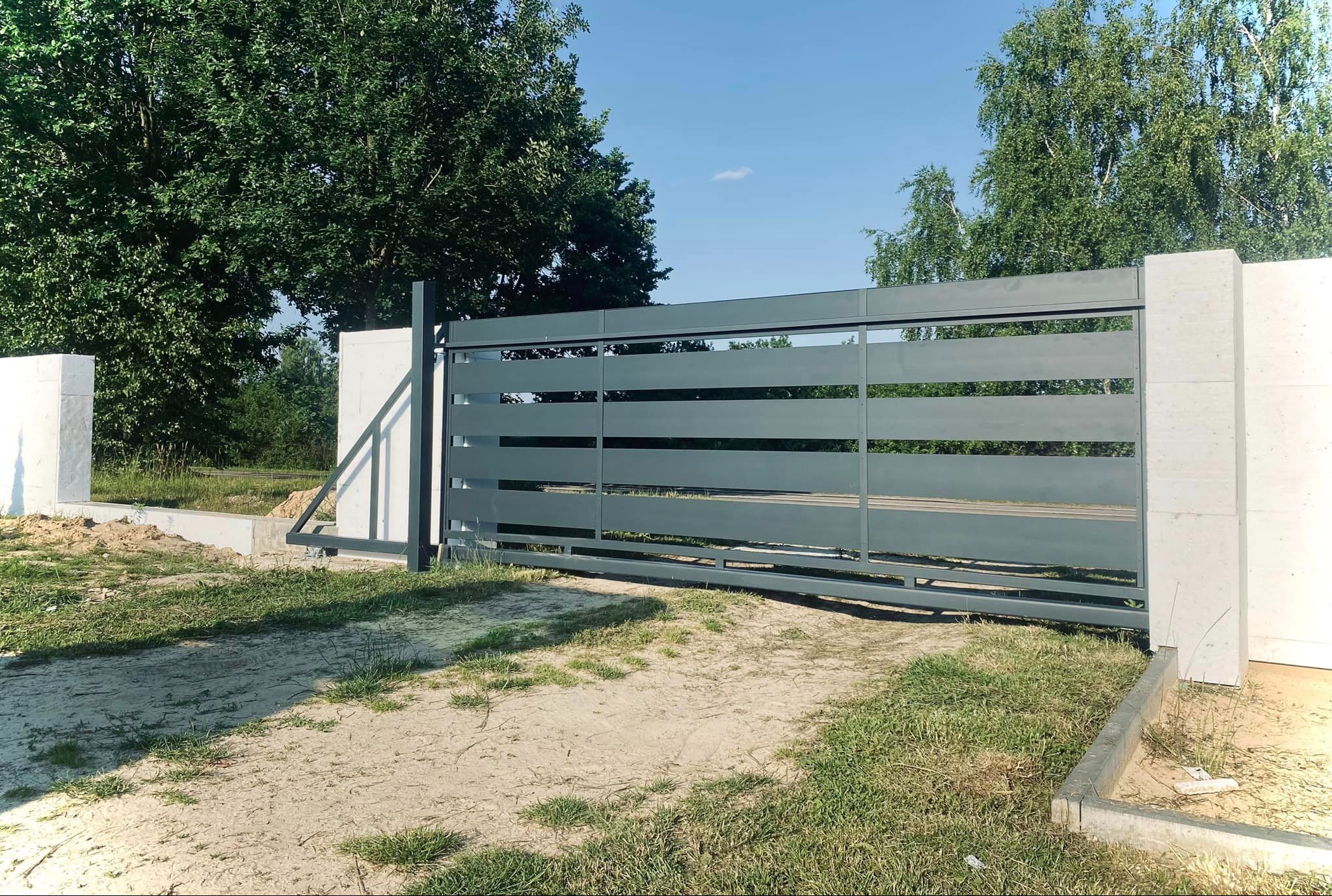 Nowoczesna, przesuwna brama wjazdowa w kolorze grafitowym, z poziomymi panelami, zamontowana na betonowych słupkach. Widoczne zielone drzewa i błękitne niebo w tle.