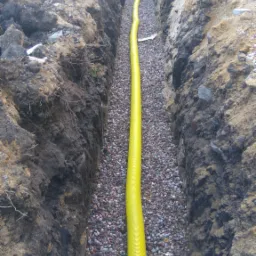 Żółta rura drenażowa ułożona w wykopie wypełnionym drobnymi kamieniami, widoczne ściany wykopu z ziemi i piasku, w tle budynek w trakcie budowy i pracownik.