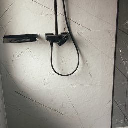 Wykończona łazienka z czarną armaturą: deszczownica, bateria i półka. Białe płytki imitujące marmur z szarymi żyłami. Uwagę przykuwa odpływ liniowy w podłodze.