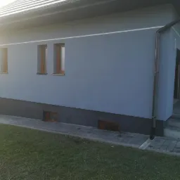 Świeżo wykończona ściana domu w odcieniach szarości z widocznymi oknami i rynnami, z fragmentem trawnika w słonecznym świetle