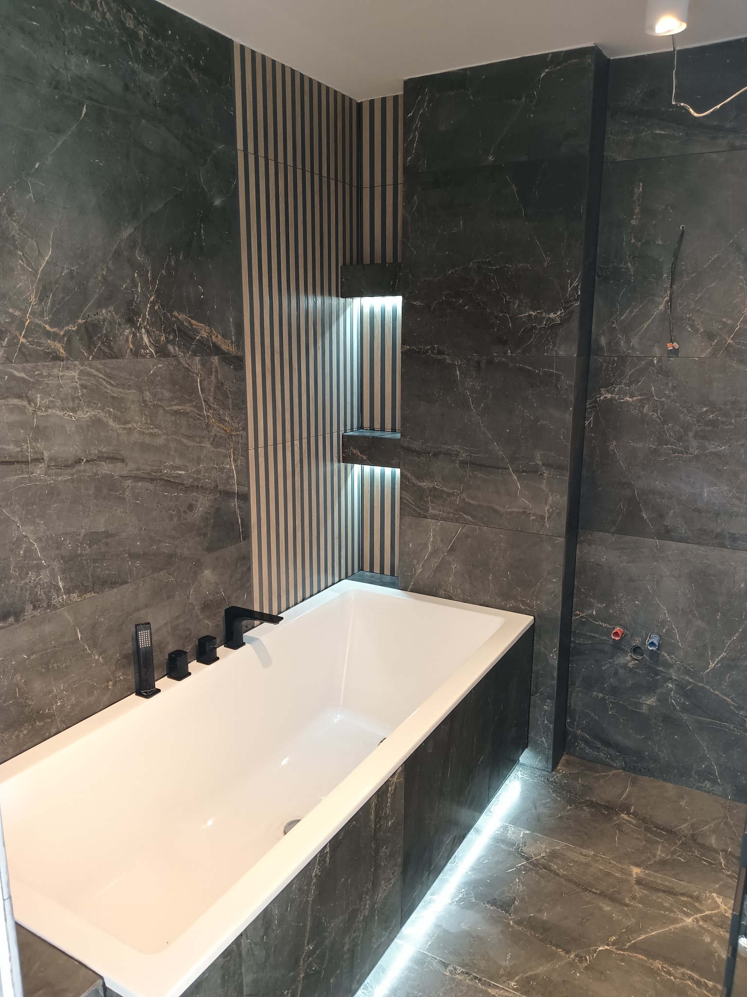 Nowoczesna łazienka z wanną i marmurowymi płytkami w odcieniach szarości, oświetlenie LED pod wanną i w niszach ściennych, czarna armatura.