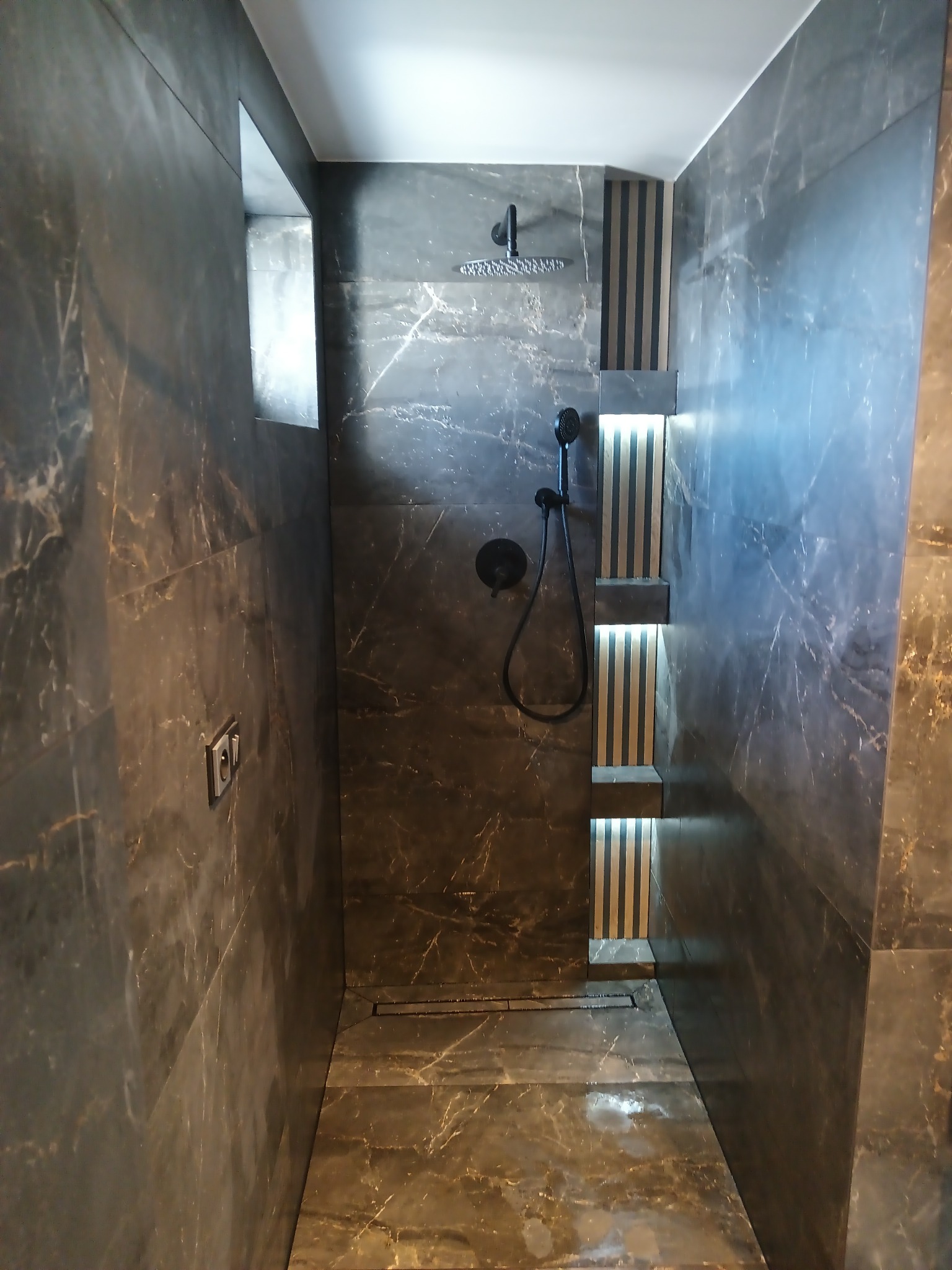 Kabina prysznicowa wykończona ciemnym marmurem, z wbudowanymi półkami podświetlonymi paskami LED i deszczownicą.