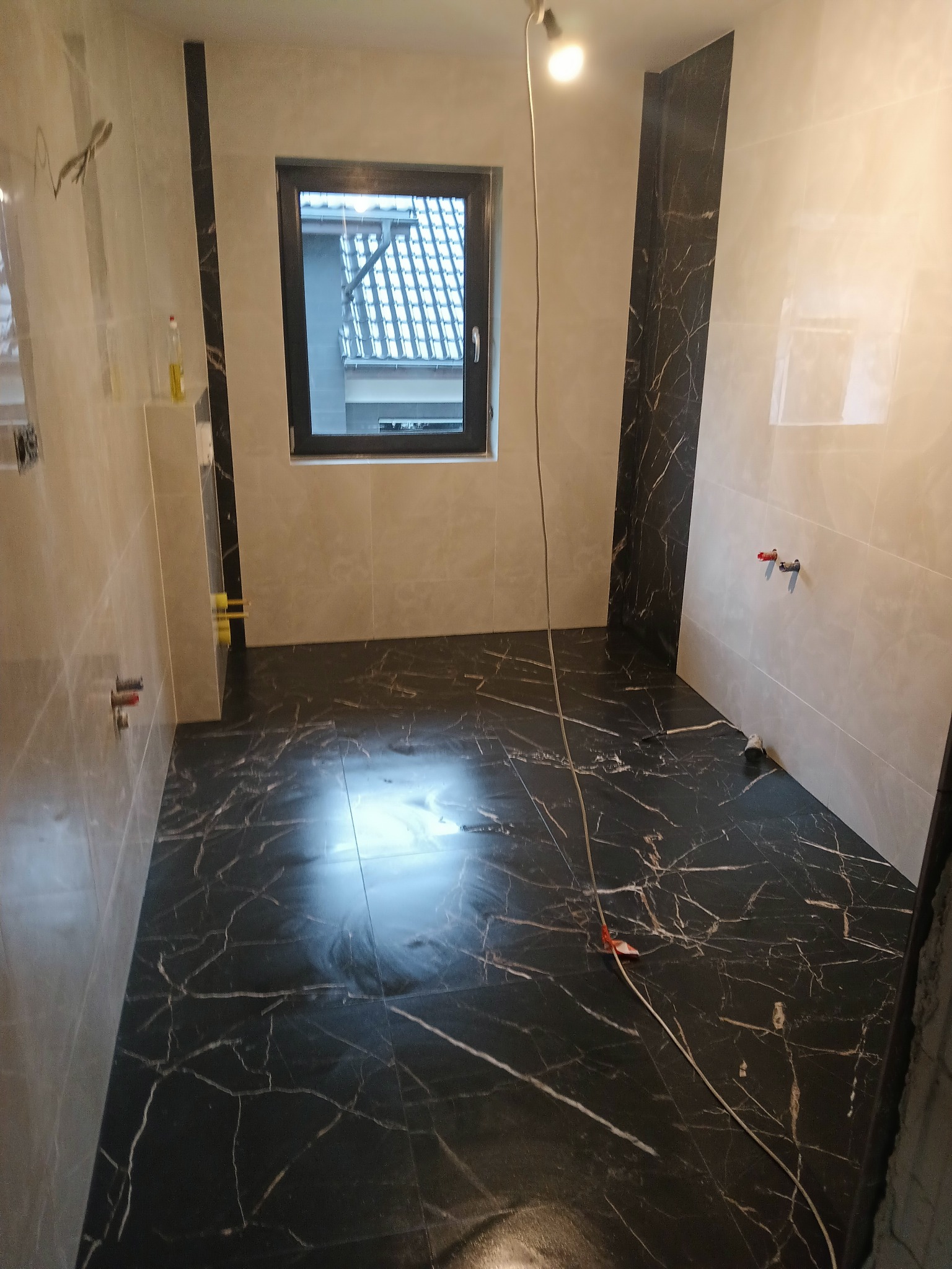 Łazienka w trakcie remontu: ułożona czarna glazura z białym wzorem na podłodze i fragmentach ścian, widoczne instalacje wodne, okno z widokiem na dach.