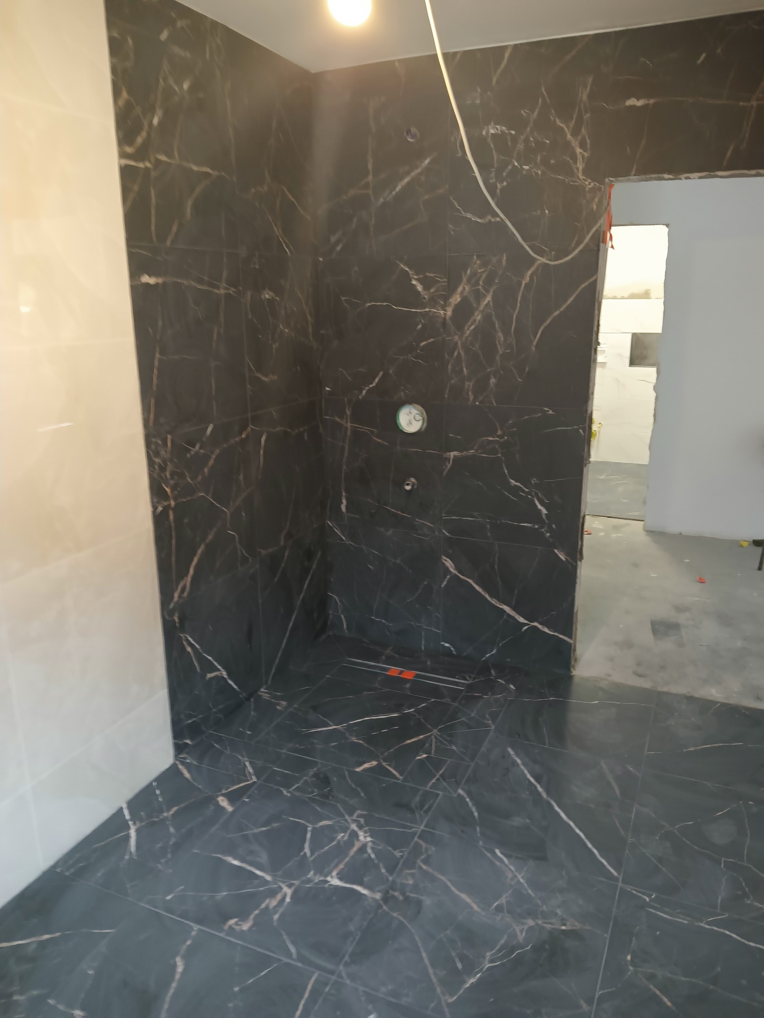 Wykończona kabina prysznicowa z czarnych płytek imitujących marmur, z widocznym odpływem liniowym i fragmentem ściany wyłożonej jasnymi płytkami.